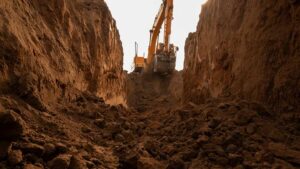 Excavator shaping a residential worksite beside a deep trench, exposed soil layers, gravel, and safety barriers.