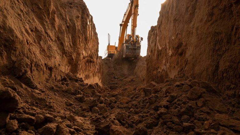 Excavator shaping a residential worksite beside a deep trench, exposed soil layers, gravel, and safety barriers.