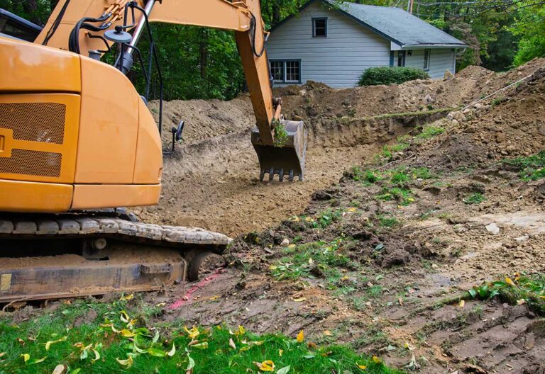 Marked excavation area beside a house with utility flags, cleared lawn, and protected landscaping. KEYWORDS: ["residential yard marked for excavation", "underground utility flags and paint markings", "mini excavator beside house", "cleared lawn prepared for digging", "protected tree roots near trench", "excavated soil pile and drainage trench", "temporary ground protection mats", "residential foundation excavation", "yard grading and backfilling"]