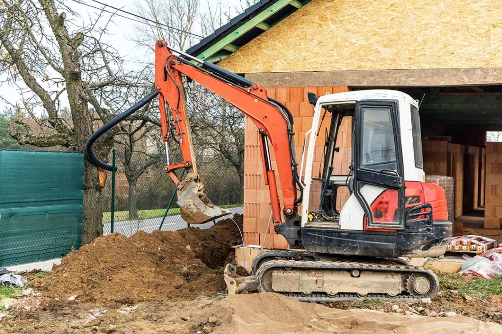 Excavator preparing a level foundation area beside a residential construction site with soil and gravel visible.